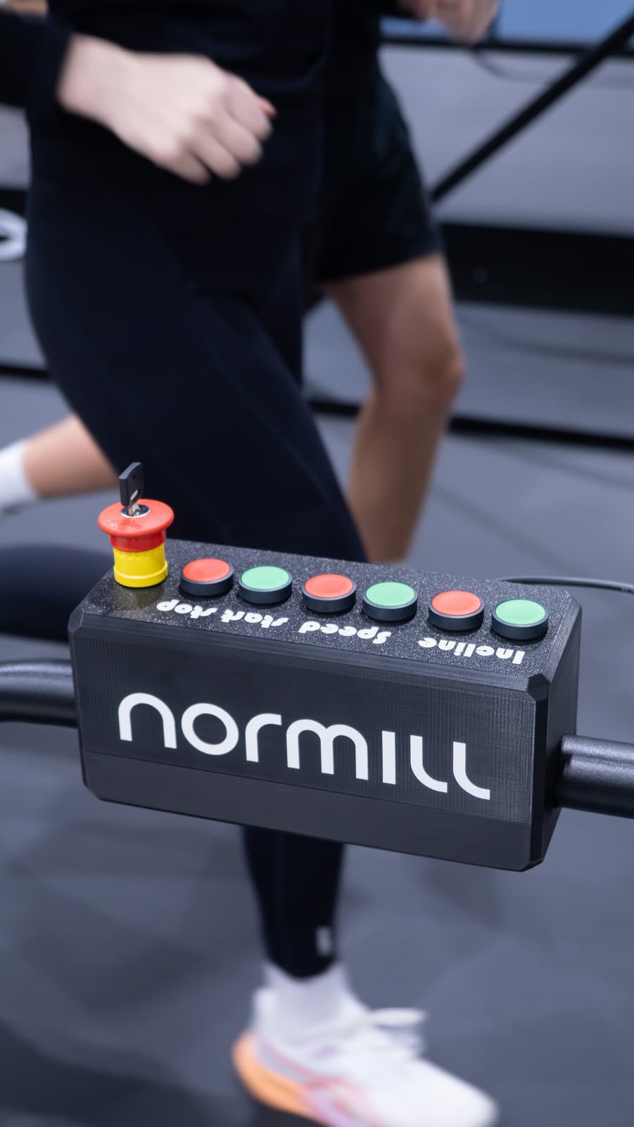 Handlebar control box marked Incline, Speed, Start and Stop, with a key-release emergency stop, while someone trains behind it.