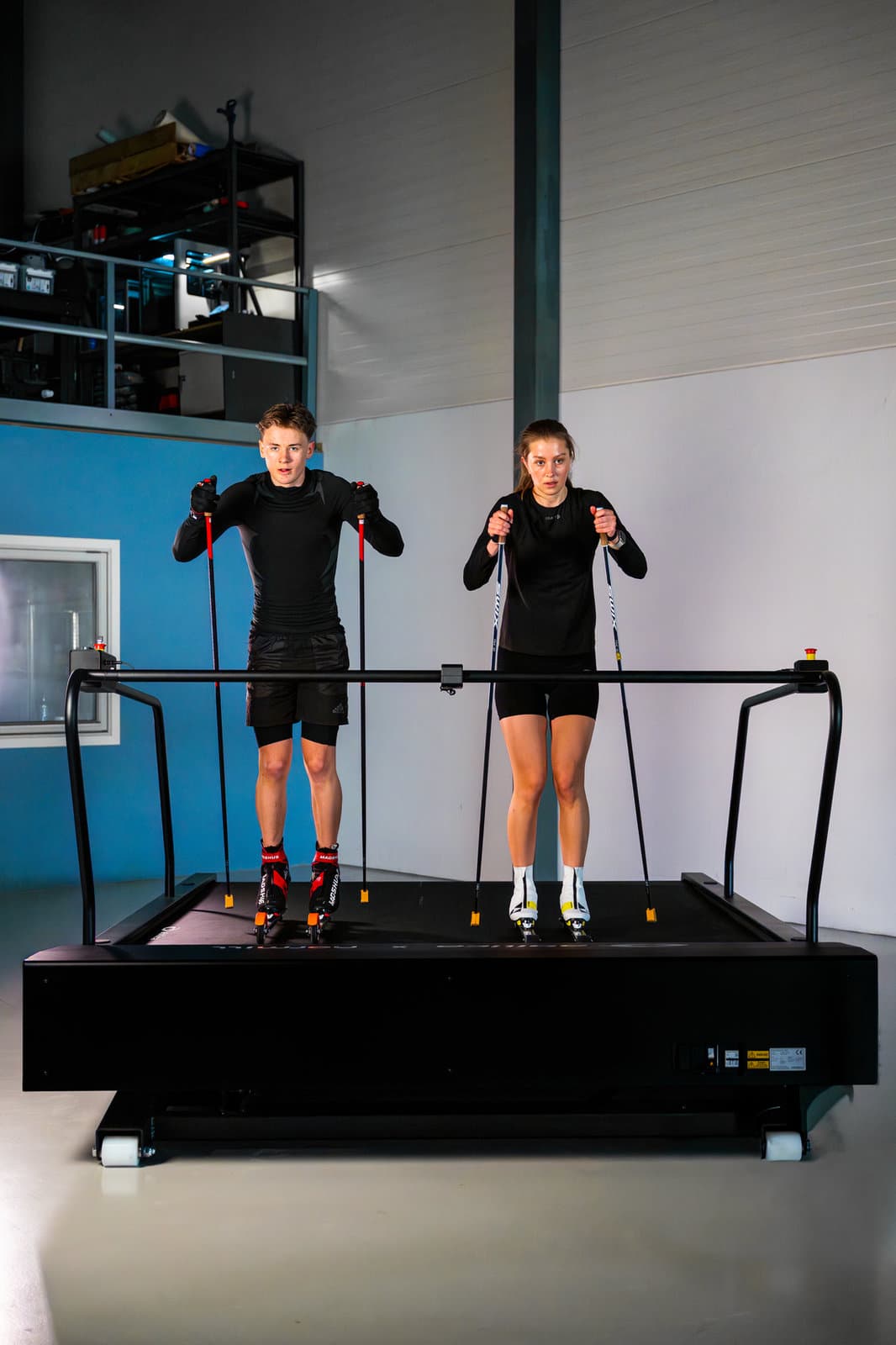 Two skiers training side by side on roller skis on a wide Normill treadmill.