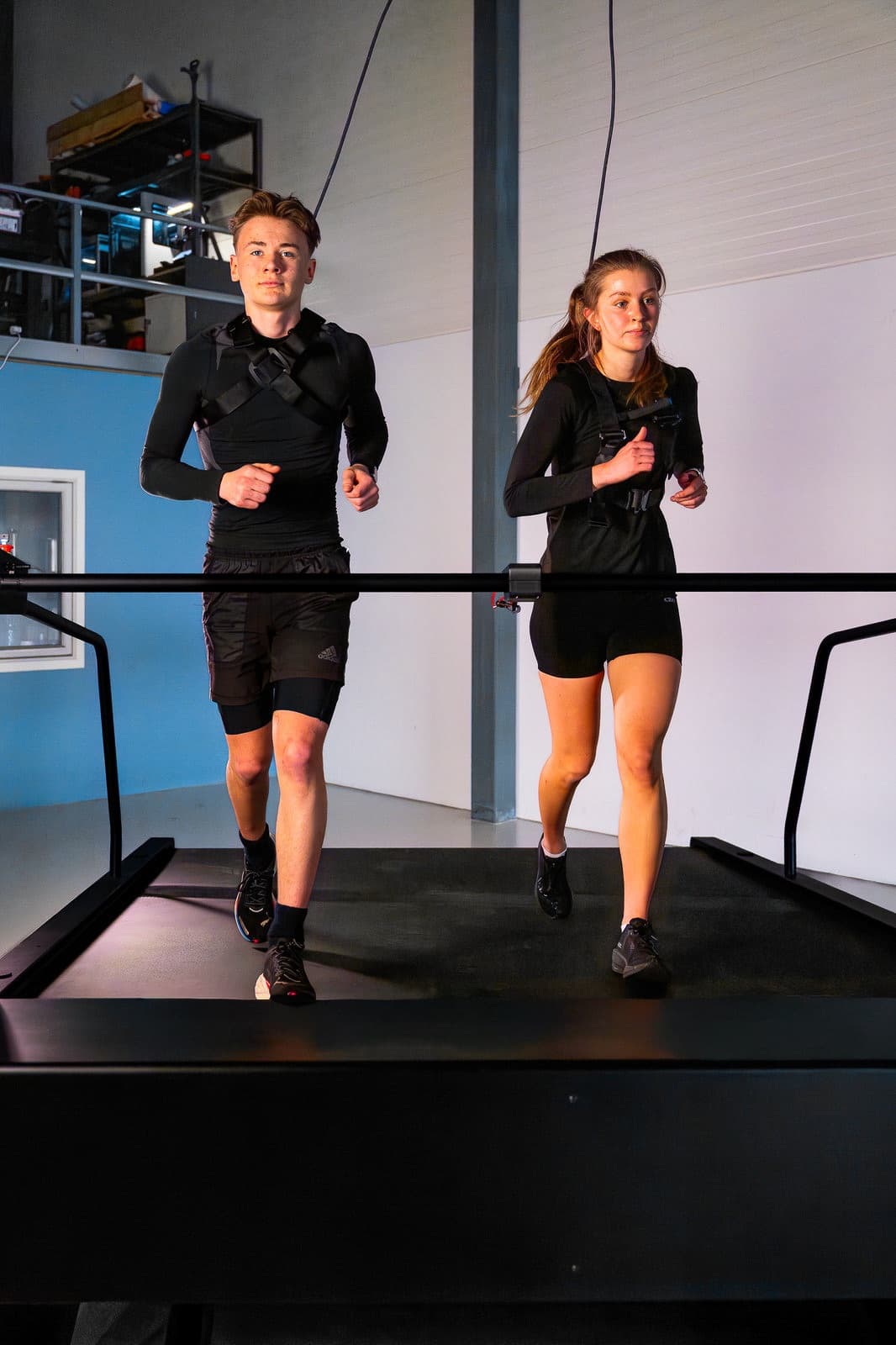 Two runners in safety harnesses running side by side on a Normill treadmill.