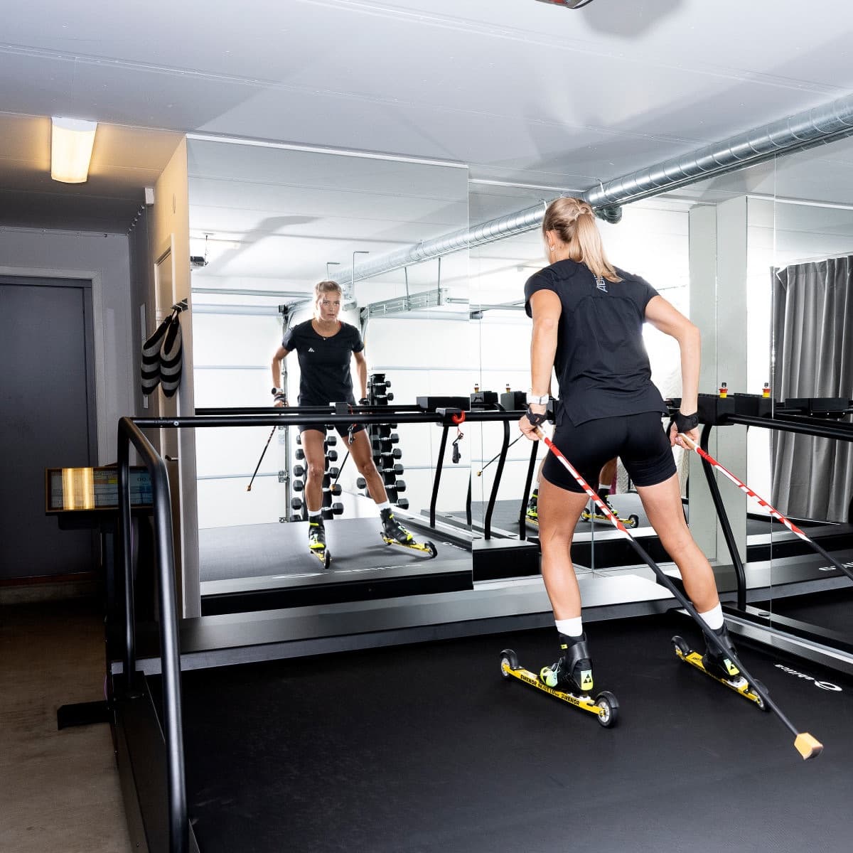A skier on roller skis seen from behind on a Normill treadmill in a mirrored training room.